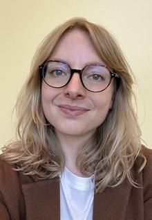 A blonde white woman in brown round glasses and a brown cardigan fit for a librarian.